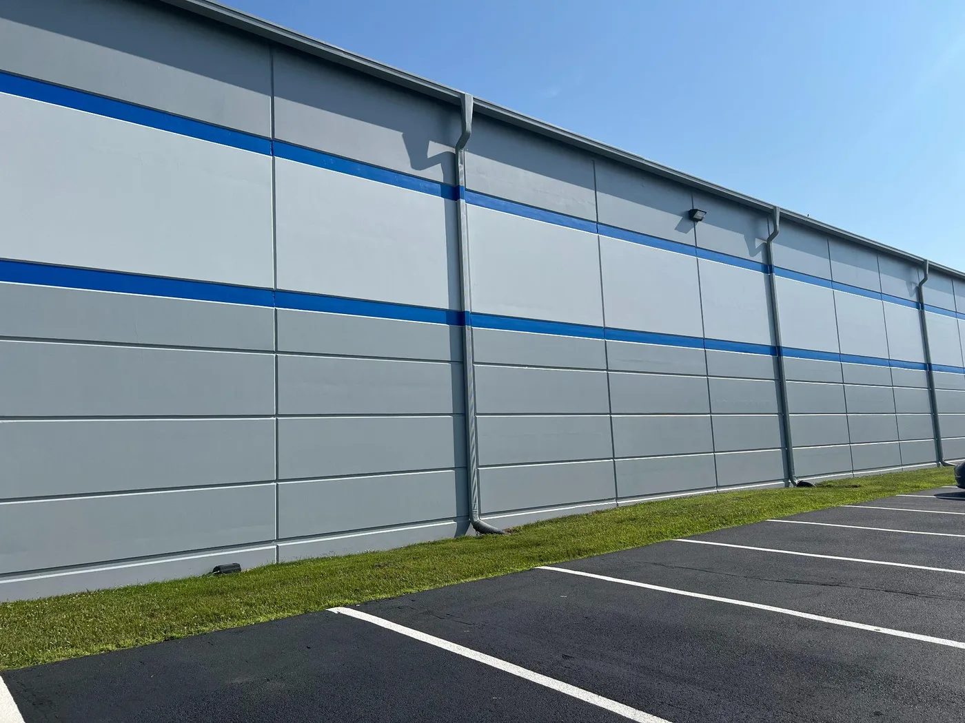 Long metal warehouse wall painted light grey with a blue horizontal stripe, beside a striped parking lot