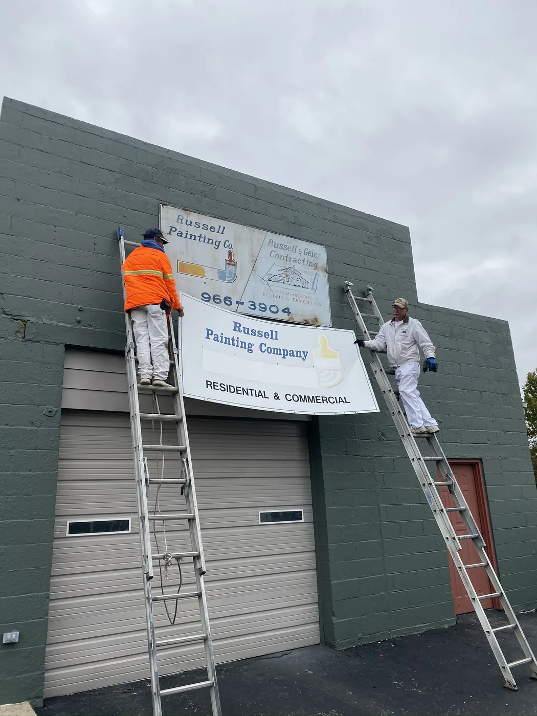 Two Russell Painting Co painters on extension ladders painting the exterior of a commercial building dark green